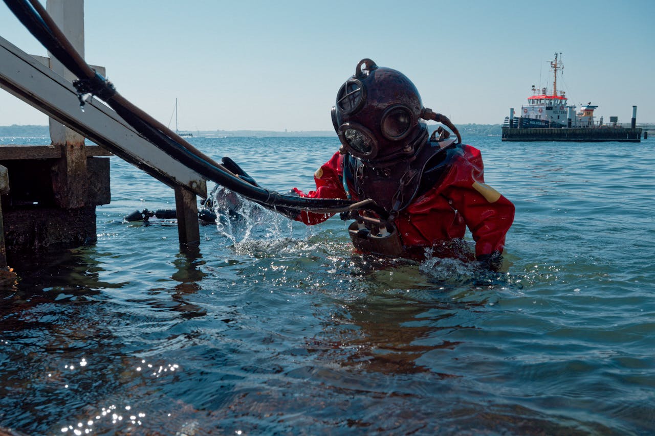 our-experience A diver in a vintage diving suit emerges from the ocean near a dock with a tugboat in the background.