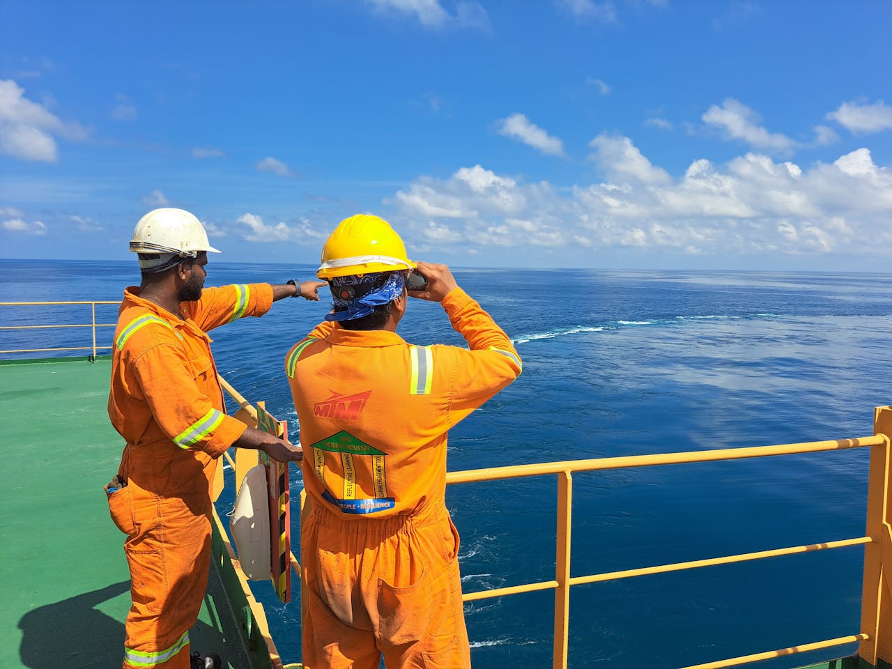 ours-journey Two workers on an oil rig deck wearing safety gear inspect the ocean horizon under a clear sky.